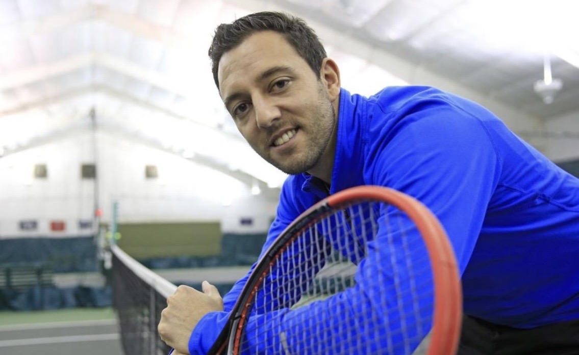 Mike Manzella leaning on a tennis net inside an indoor racquet facility